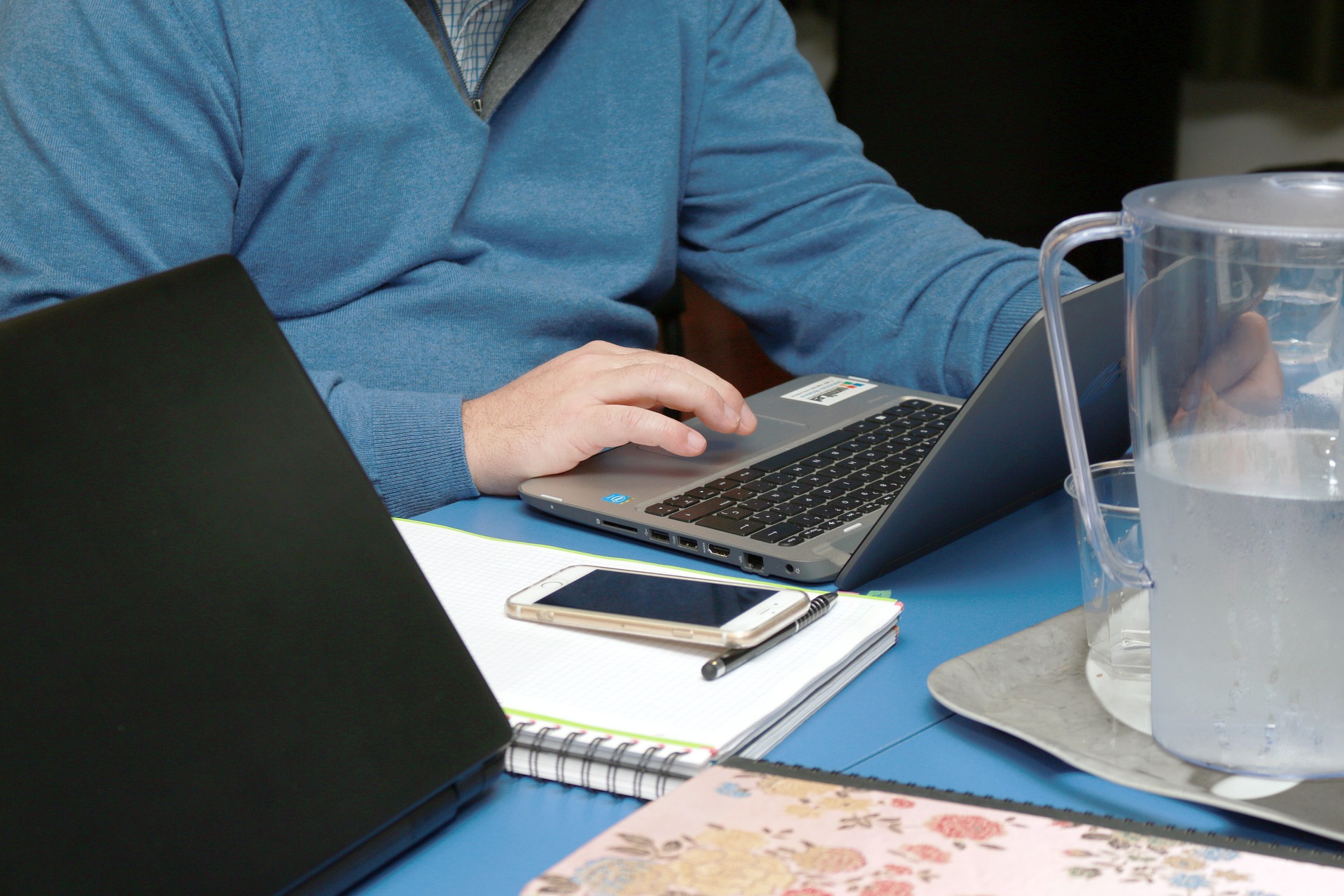 Un hombre escribiendo en un portatil, al lado esta un celular y varios vasos con agua