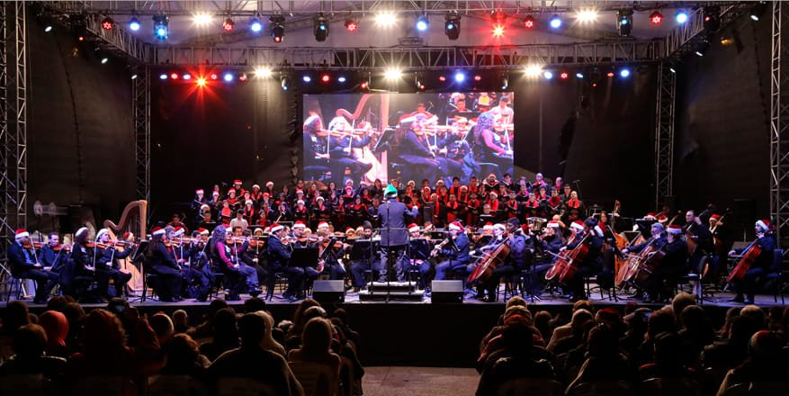 Un escenario, donde están varias personas tocando instrumentos musicales con un publico de espaldas 