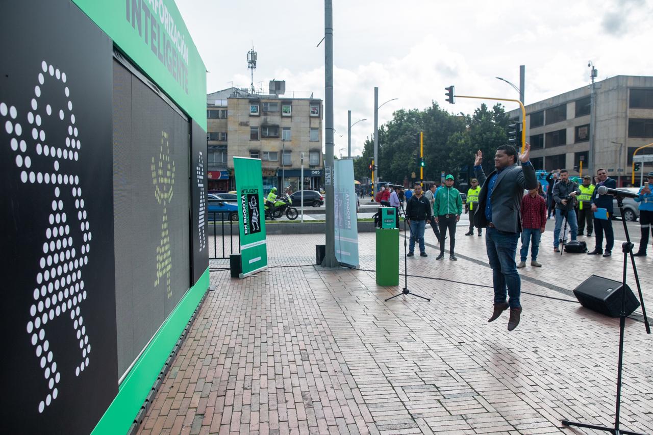 En un evento publico sobre la semaforización en la ciudad, un hombre participa en un juego en la mitad del evento, saltando enfrente de una pantalla