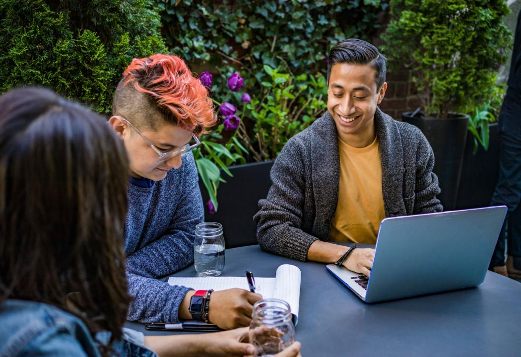 Tres jóvenes reunidos en una mesa con un portátil y varios cuaderno, hablando y riendo entre ellos 