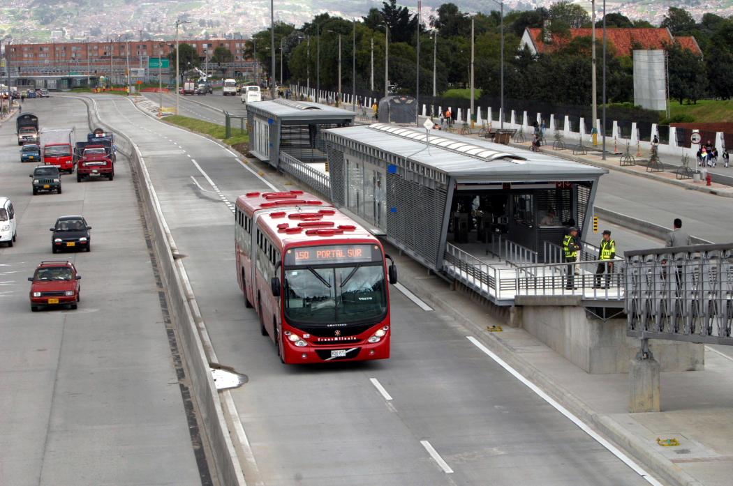 Una calle de la ciudad, un transmilenio saliendo de una estación, al lado del carril varios carros particulares