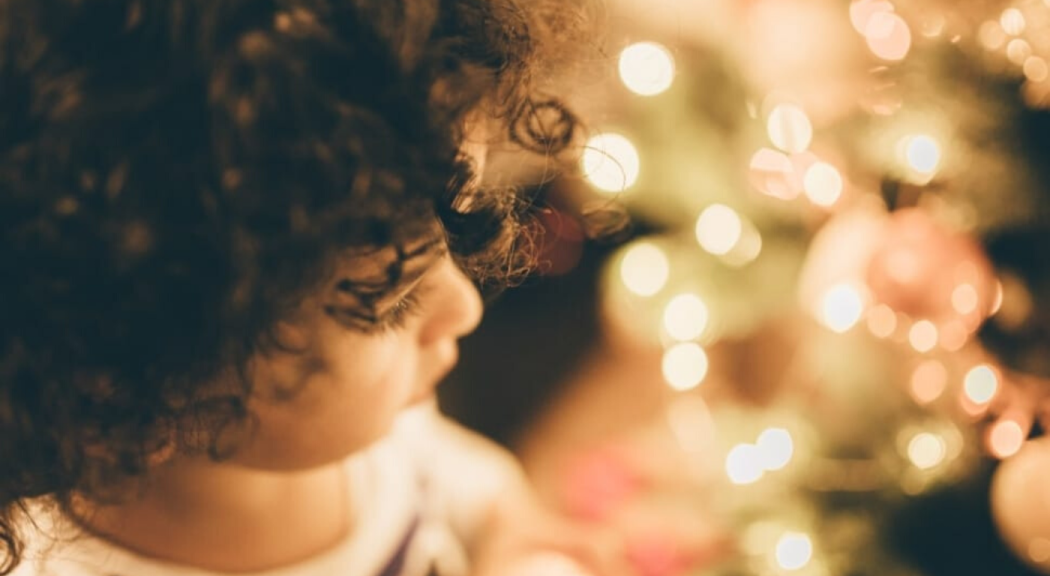 Imagen ilustrativa. Niño blanco de cabellos crespos mira un árbol de Navidad.