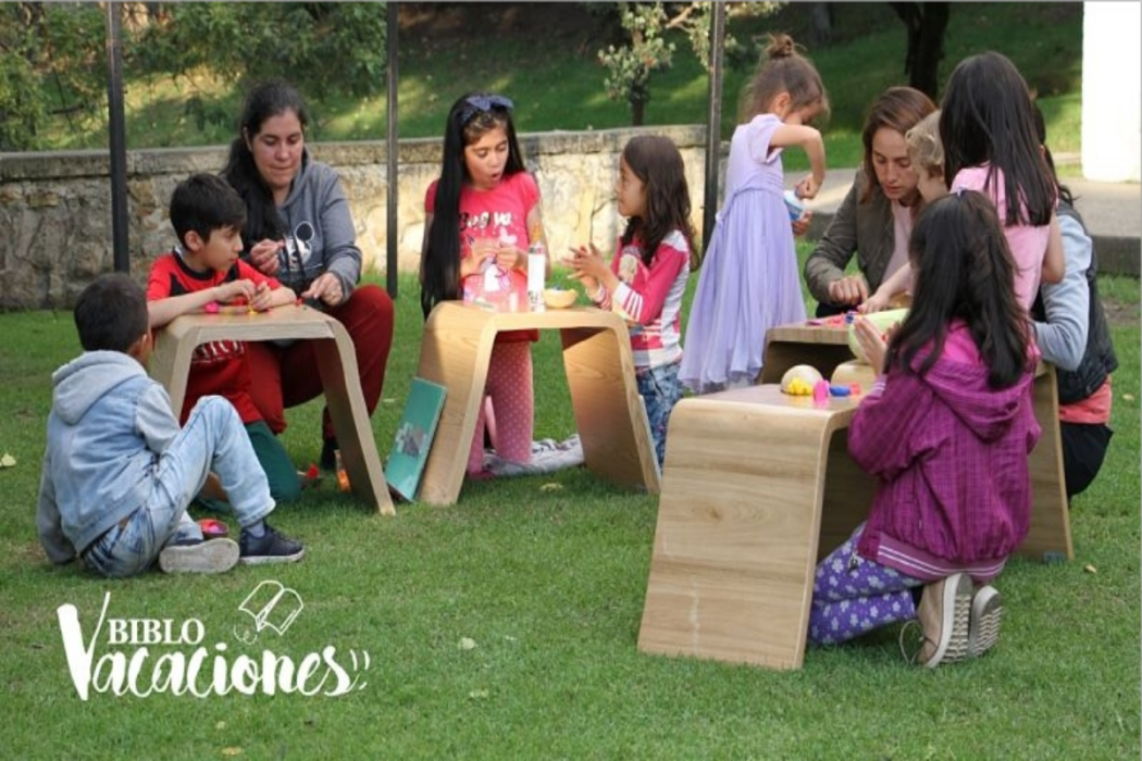 Hay varios niños sentados en el pasto, con mesas, leyendo libros