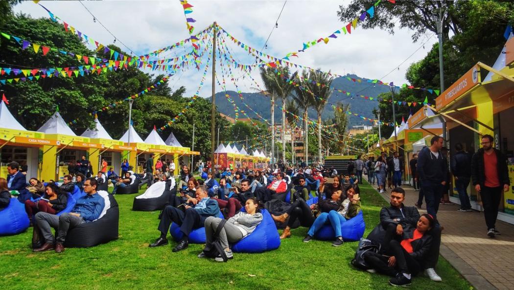 Varias personas sentadas en cojines al aire libre, alrededor muchas carpas con mercancia