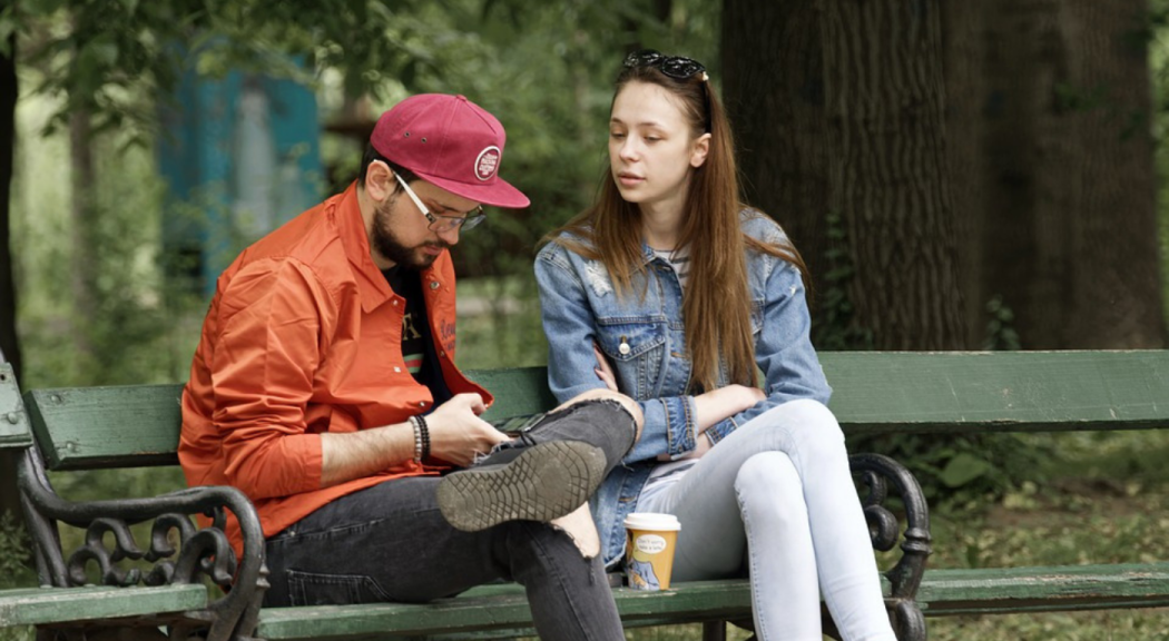 Una pareja joven se sienta en la banca de un parque a conversar.