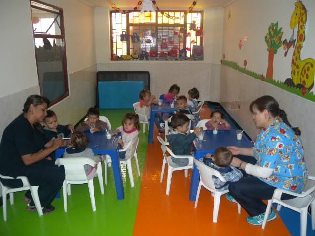 Plano general de un jardín infantil donde dos profesoras atienden a un grupo de niños pequeños.